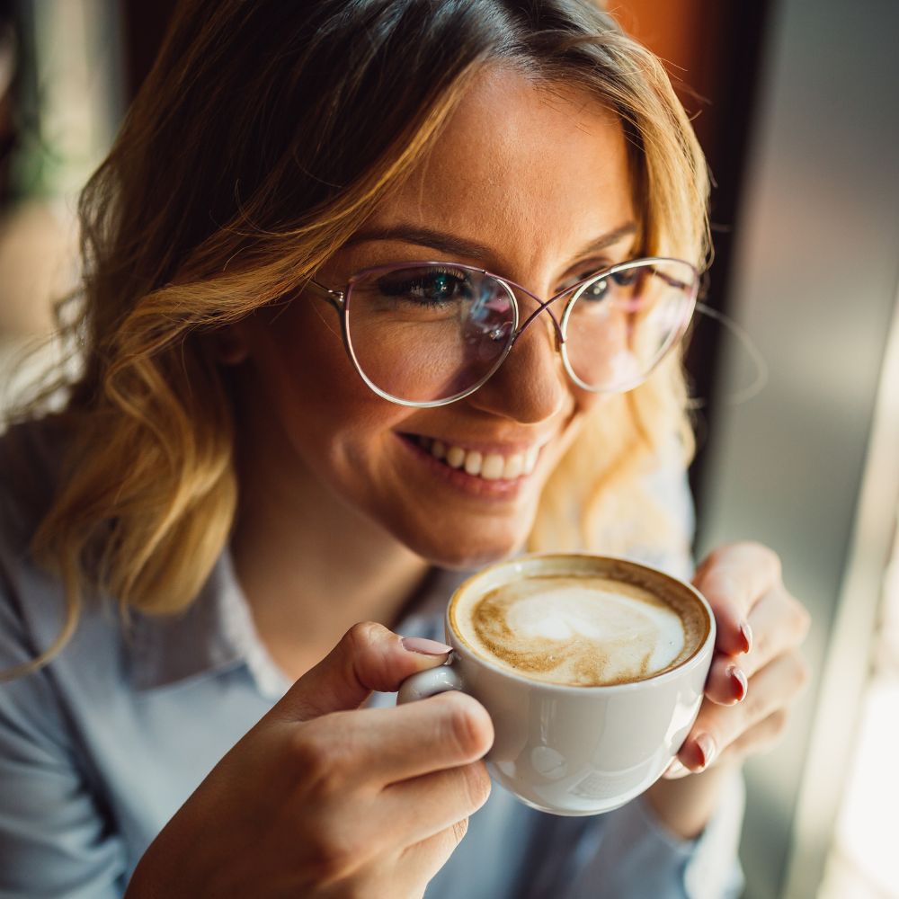 Lächelnde Frau mit Brille trinkt Cappuccino am Fenster, Nahaufnahme mit klaren Brillengläsern und warmem Licht.
