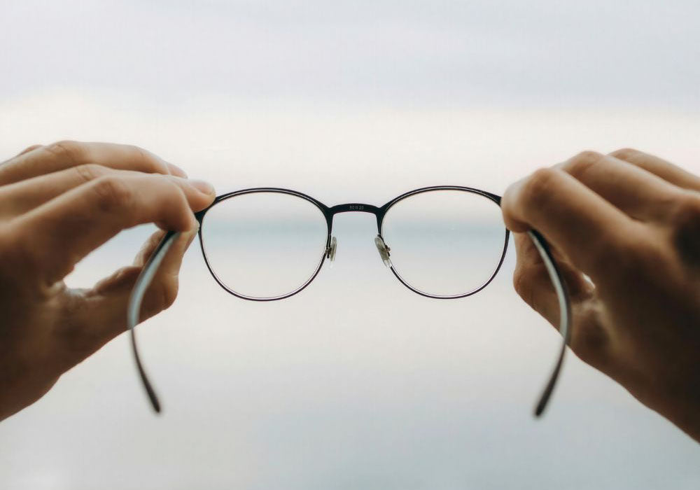 Hände halten runde Brille mit klaren Gläsern vor hellem Hintergrund, Symbol für saubere Sicht und beschlagfreie Brillen.