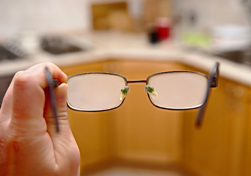 Hand hält beschlagene Brille in Küche, eingeschränkte Sicht durch Nebel auf den Gläsern im Alltag.